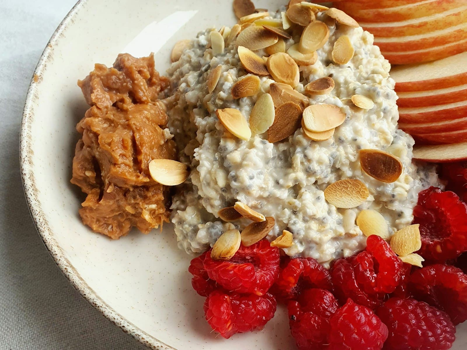Schale mit cremigen Overnight Oats mit Chiasamen, gerösteten Mandelblättchen, Apfelspalten, frischen Himbeeren und einem Klecks Nussmus