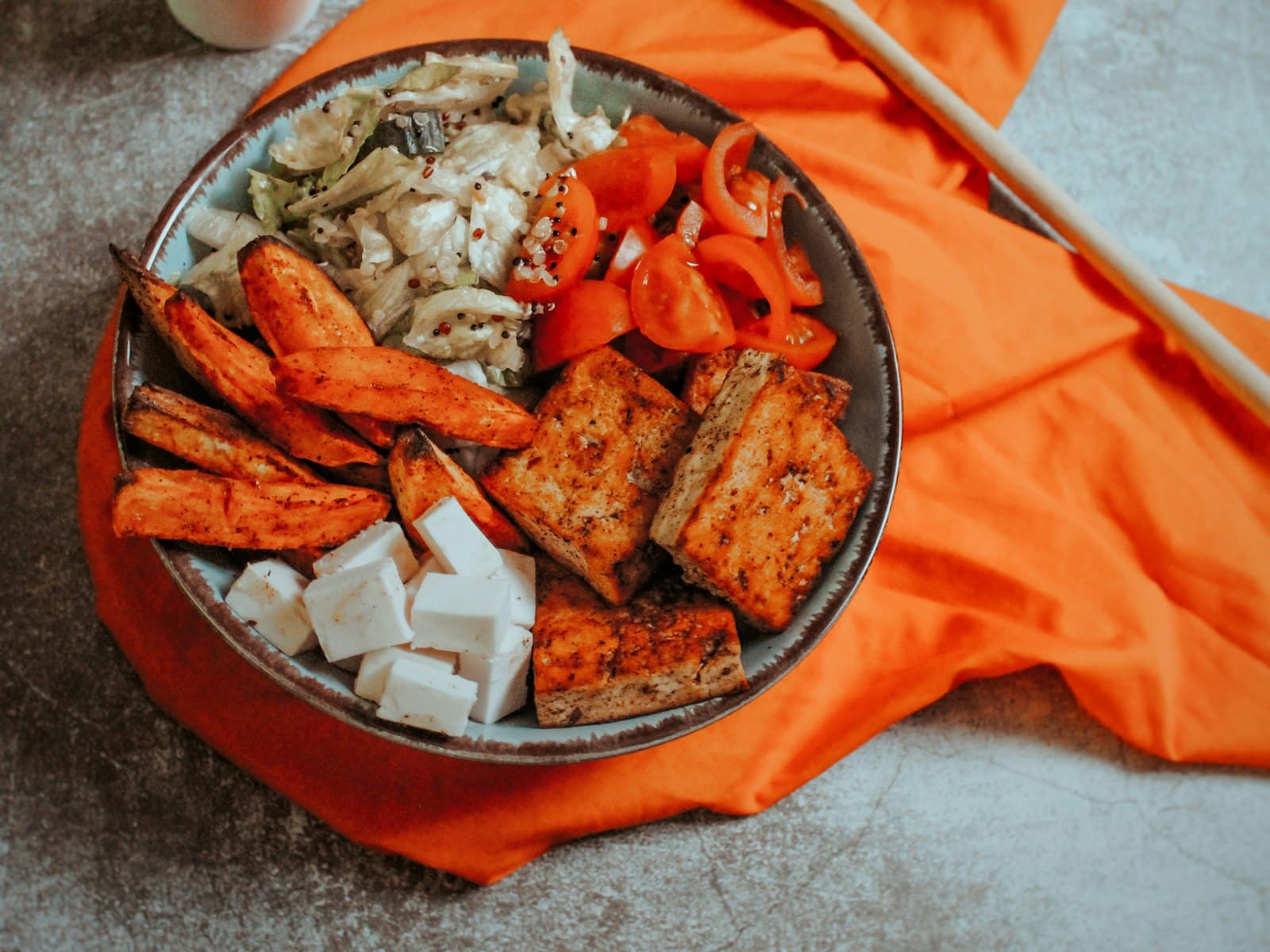 Schale mit gebratenen Tofuscheiben, orangefarbenen Ofengemüse-Spalten, Kirschtomaten, Krautsalat mit Quinoa und hellen Würfeln auf einem orangefarbenen Tuch