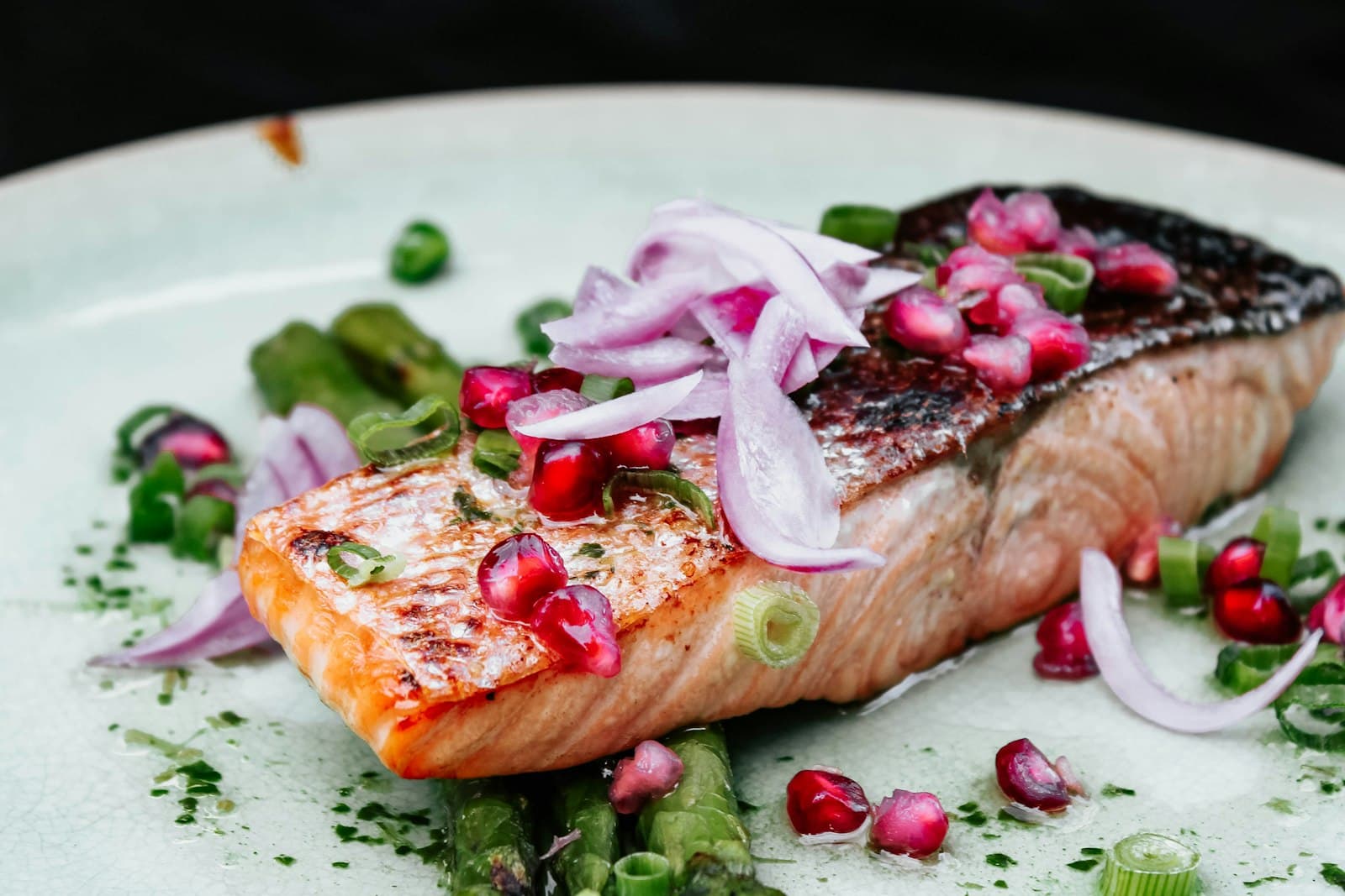 Gebratenes Lachsfilet mit knuspriger Haut auf grünem Spargel, garniert mit roten Zwiebelstreifen, Granatapfelkernen und Frühlingszwiebeln