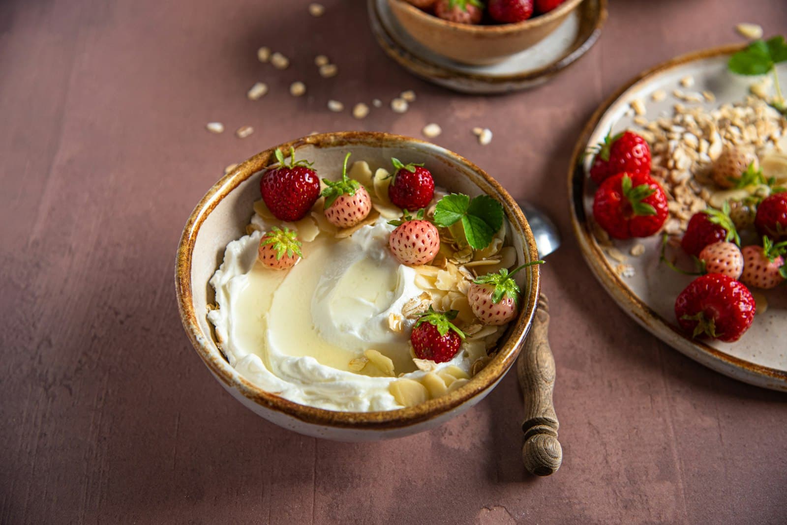 Schale mit cremigem Joghurt, Honig, Mandelblättchen und roten und weißen Erdbeeren auf rosébraunem Untergrund, daneben ein Teller mit Haferflocken und Erdbeeren